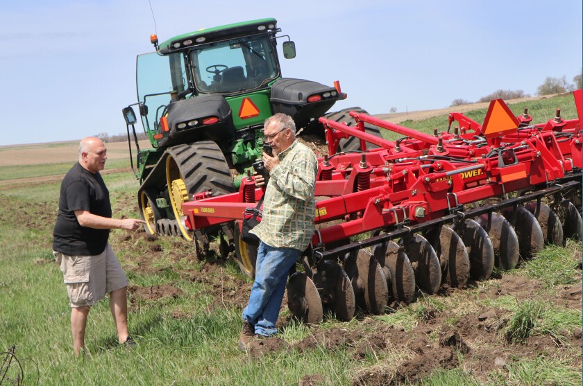 A man in shorts has delivered a lunch bucket to his father-in-law farmer, leaning on a disk.