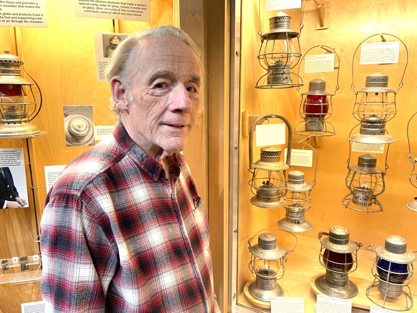 Elderly light-skinned man wearing checkered flannel shirt stands before a museum display of railroad lanterns.