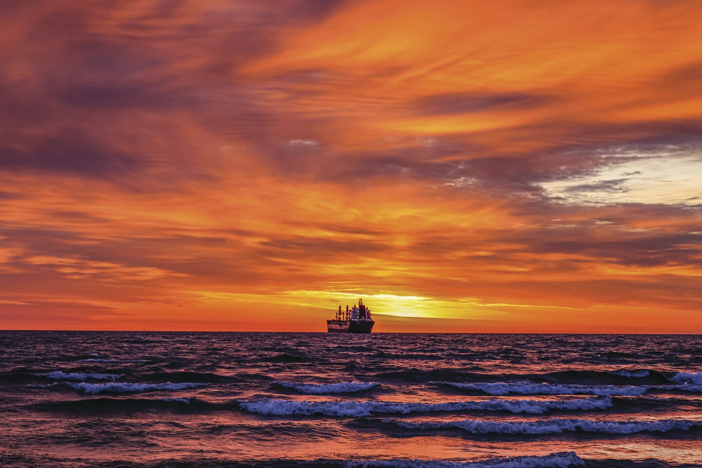 Bulk freighter sits at center on a horizon-line view of large lake, with sunrise turning sky bright orange in background.