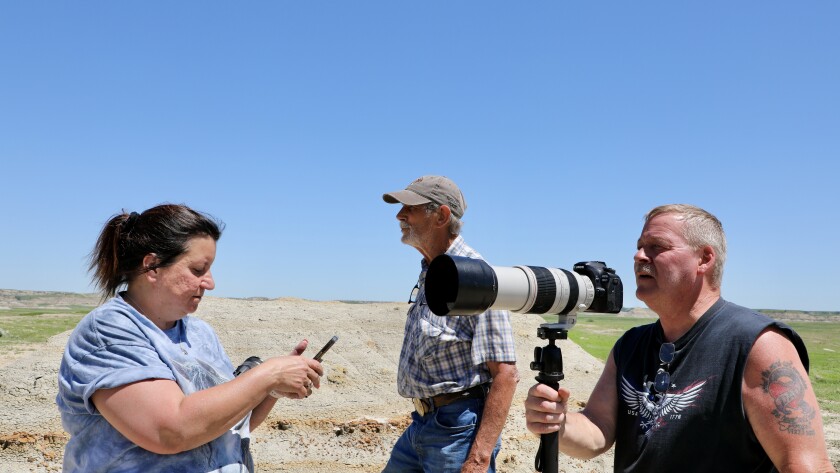 A woman uses her phone while a man in a baseball cap stares off and another man holds a camera with a long lens.