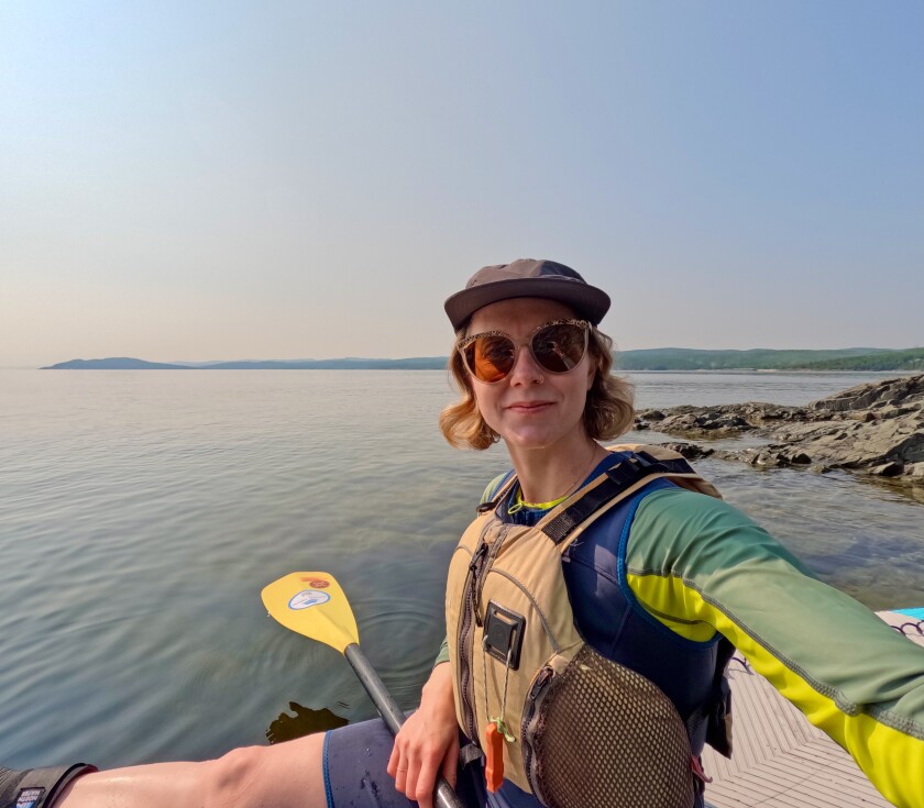 Light-skinned woman sits on a rocky lakeshore holding an oar. Wearing a lifejacket, hat, and dark glasses, she smiles slightly.
