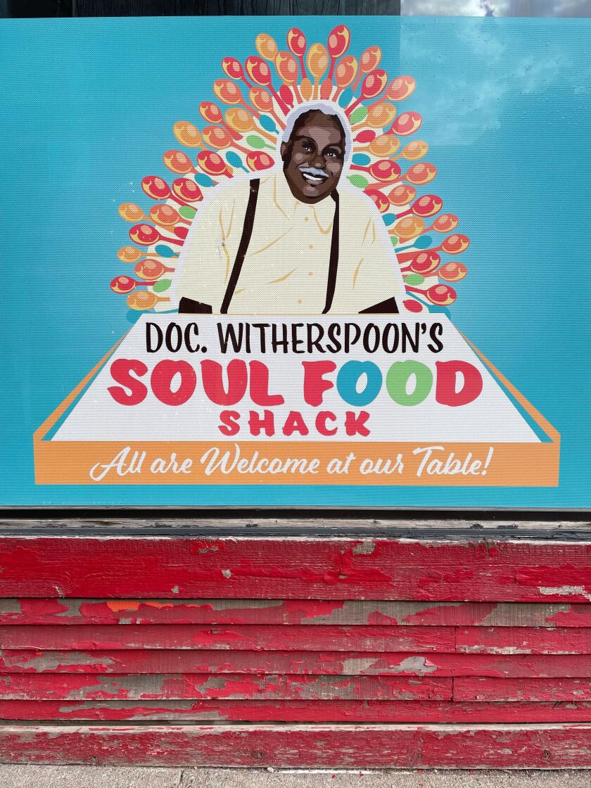 Colorful sign for Doc Witherspoon's Soul Food Shack with image of smiling Black man and the words, "All are welcome at our table!"