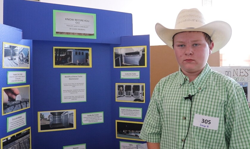A boy in a green plaid shirt and cowboy hat stands next to an educational display on trailer safety.