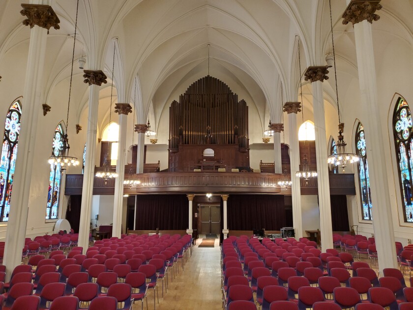 Interior view of church sanctuary with red-backed chairs replacing pews for seating. the brown pipes of a grand organ are visible in a choir loft.