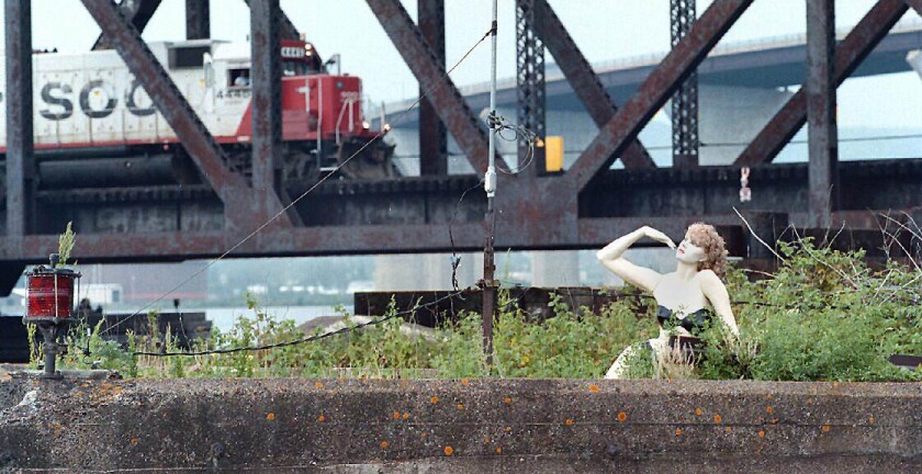 A mannequin poses beneath a bridge as a trains passes over.
