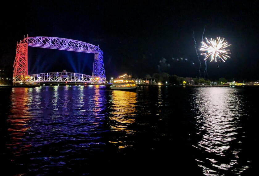 A nighttime view of Duluth Harbor shows, from left to right: the Aerial Lift Bridge, illuminated in multiple colors; a Vista Fleet boat; and fireworks in the sky.