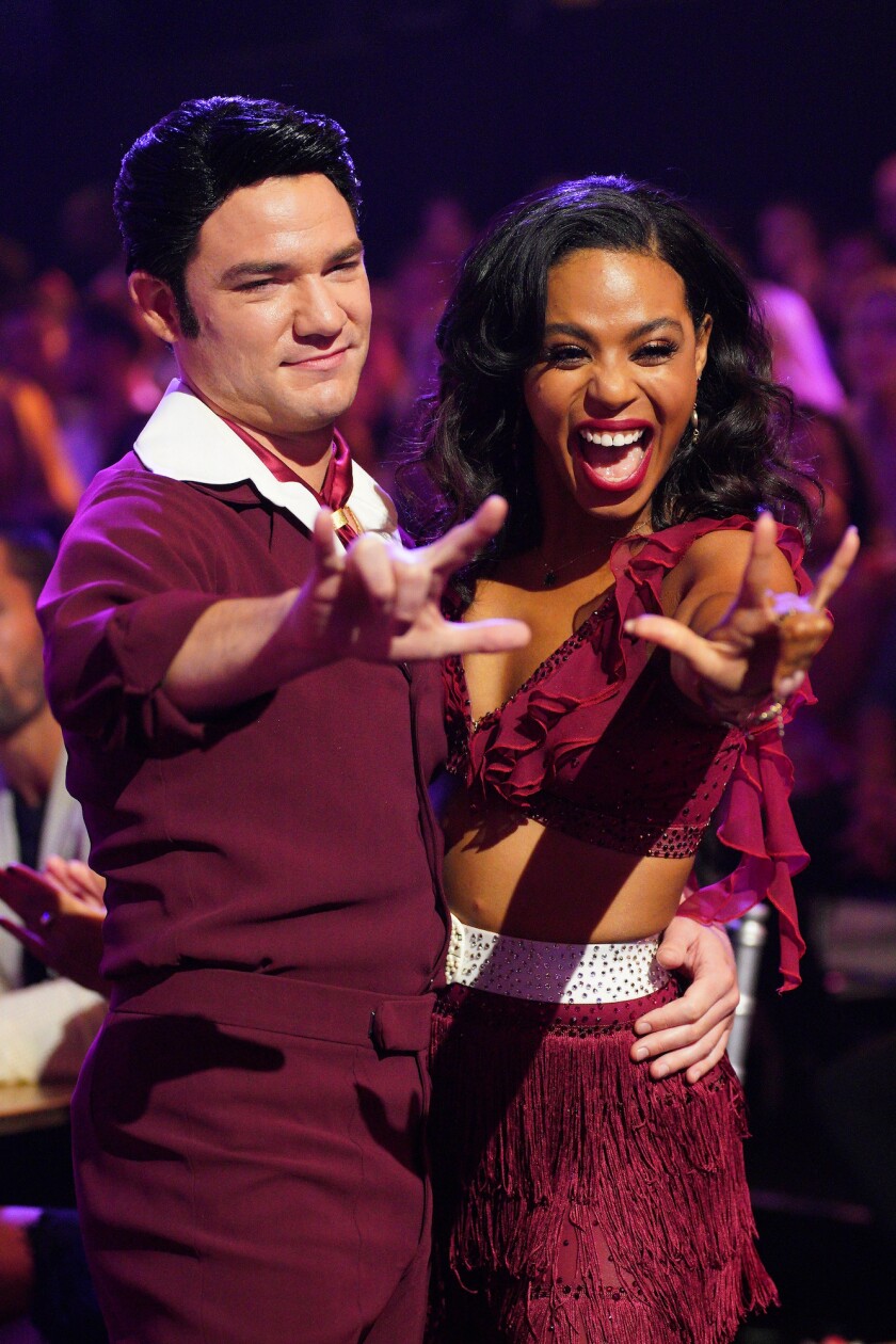 A man and woman in plum dance clothes stand with their arms around one another, making the ASL sign for love.