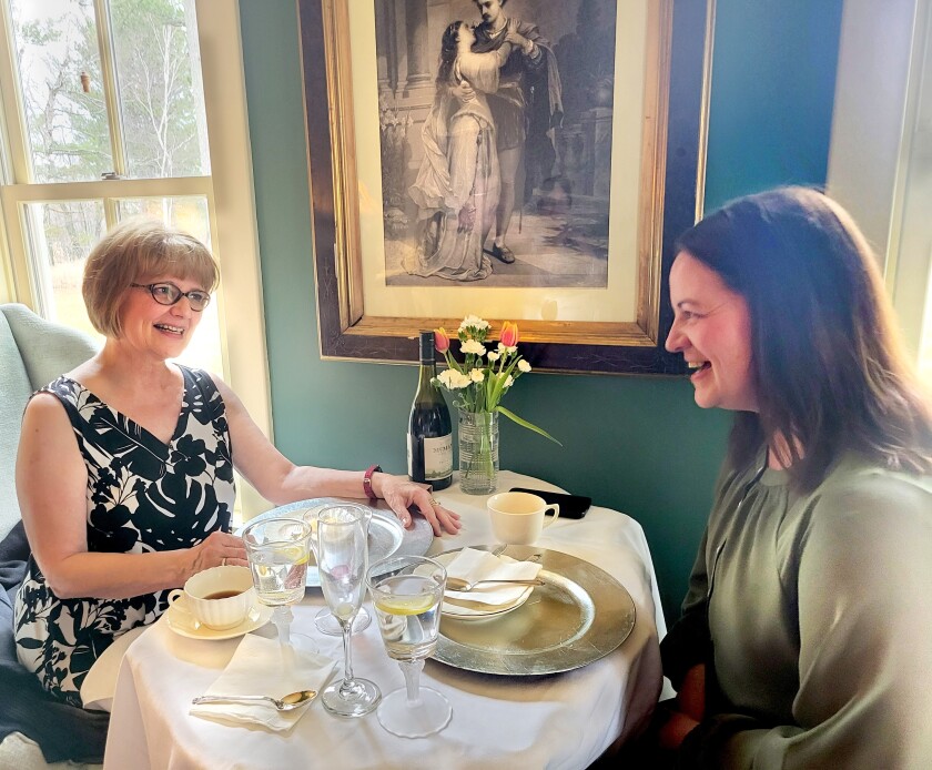 Lori Bergstedt sits across from her daughter Katy Bergstedt. Their table is covered with plates, glasses, coffee cups, a bottle of wine and flowers in a vase. The walls are a deep green, and there's an old-timey photo of Romeo and Juliet on the wall.
