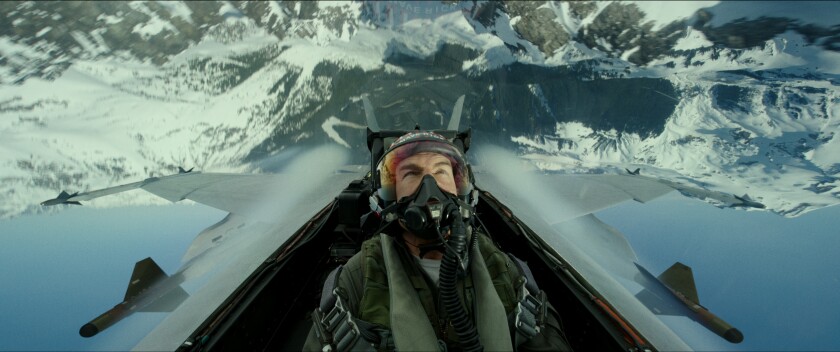 Tom Cruise, in character as "Maverick," sits in the cockpit of a fighter jet inverted over a snowy mountainscape.