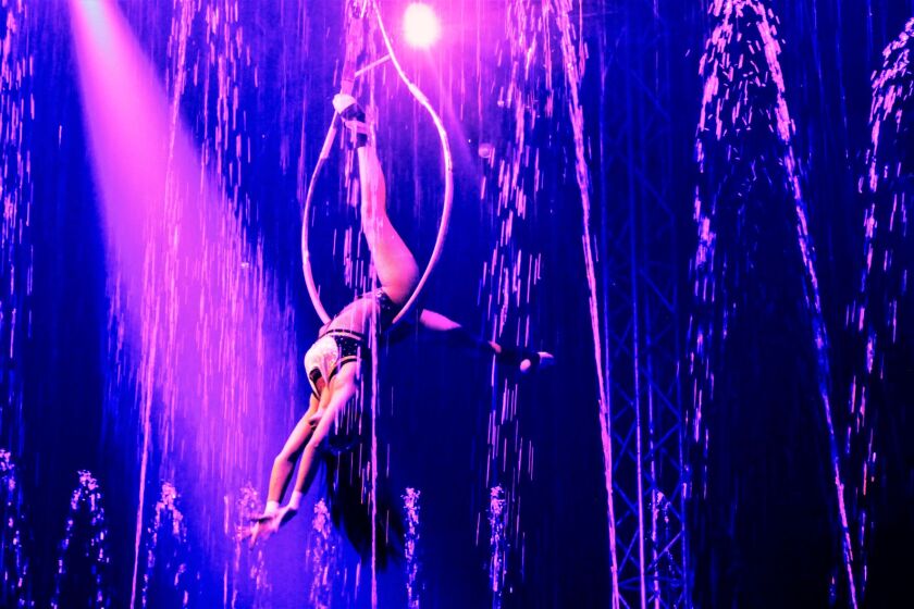 Costumed woman leans backward in aerial hoop as water fountains spurt all around her in a purple-lit circus act.
