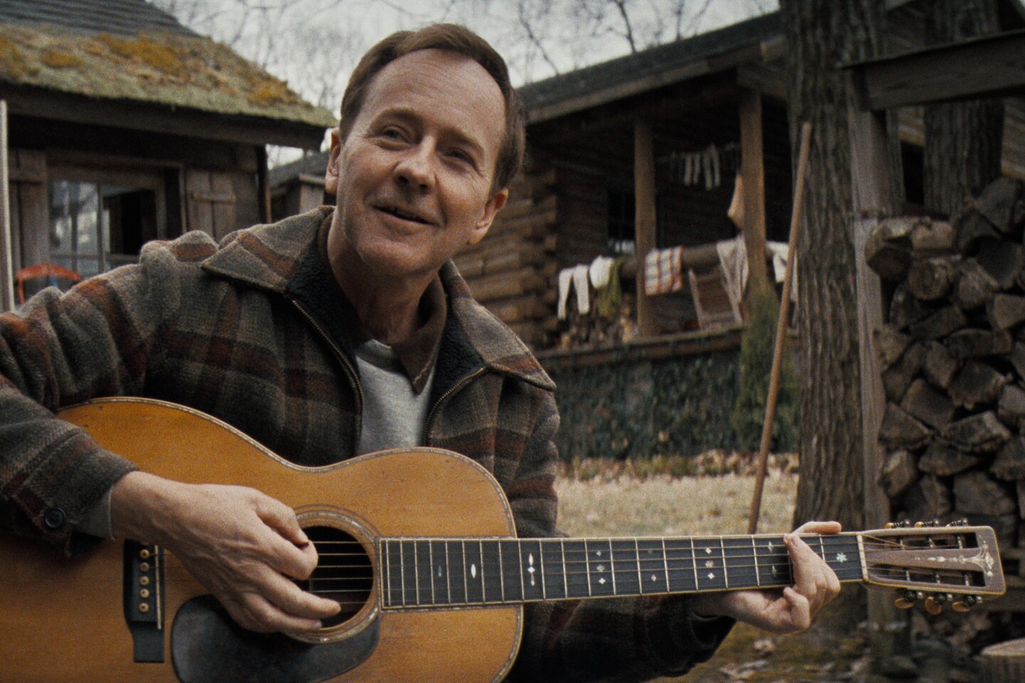 White man in his 50s, gently smiling, plays acoustic guitar near rustic wooden buildings in an outdoor setting. He wears a plaid jacket.