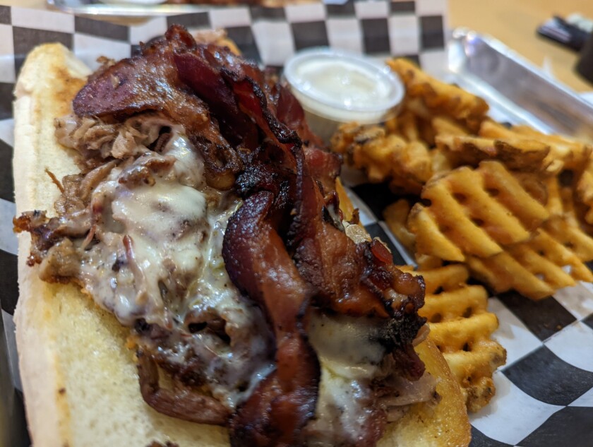 Stacks of bacon- and cheese-covered brisket sit on a long white split bun, alongside a handful of waffle fries on a piece of black-and-white checkered paper.
