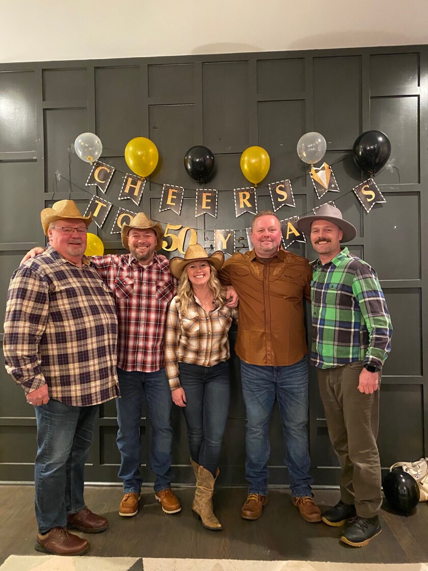 Joel Bigelow and his three sons and one daughter stand in flannel shirts in front of a sign that reads "Cheers to 50 years."