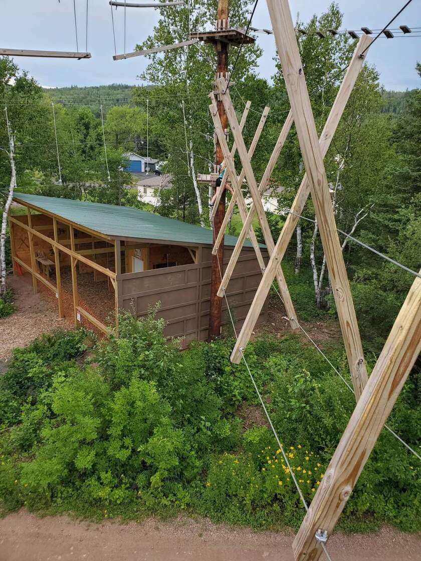 An aerial photo of a small wooden building containing ax throwing lanes. A rope course with various obstacles is visible above.