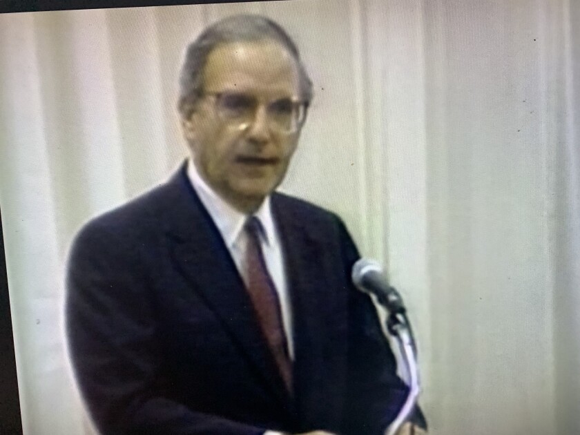 A man in a dark suit and glasses speaks in front of a microphone.