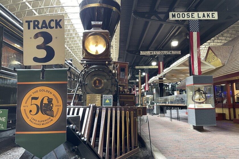 Steam locomotive with large cow-catcher and bright light stands in railroad museum. In foreground is a sign designating "3," with a golden "50" seal banner affixed.