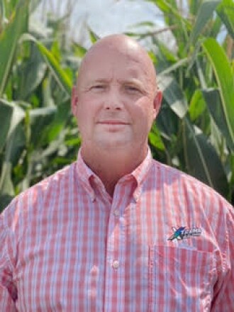 A balding farmer in a red-and-white plaid shirt stands in front of corn that is much taller.