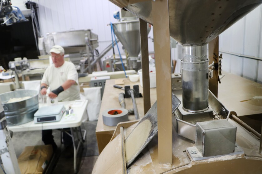 A stainless steel hammer mill stands at right, while a flour miller packs retail packages of pancake mix in a room full of equipment.