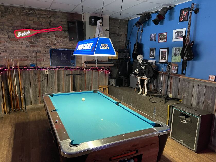 Barroom pool table with skeleton holding cigarette positioned against back wall and Bud Light fixture above table.