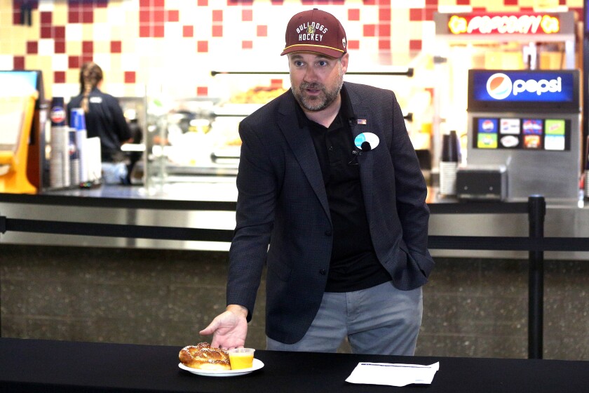 A man gestures toward a Bavarian pretzel with nacho cheese during a presentation.