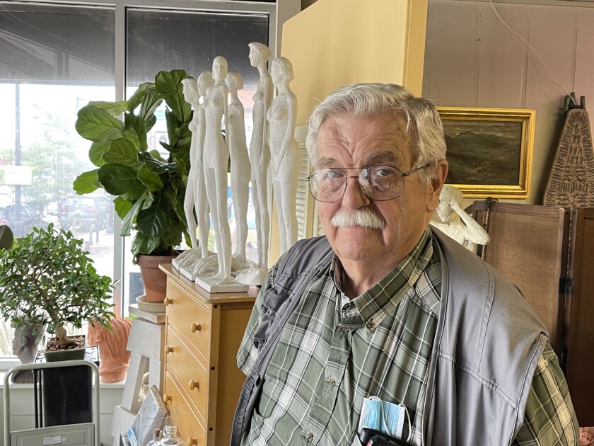 A gray-haired man wearing glasses, an olive plaid button-down shirt, and a gray waistcoat stands in front of a group of small white sculptures of stylized nude female figures.