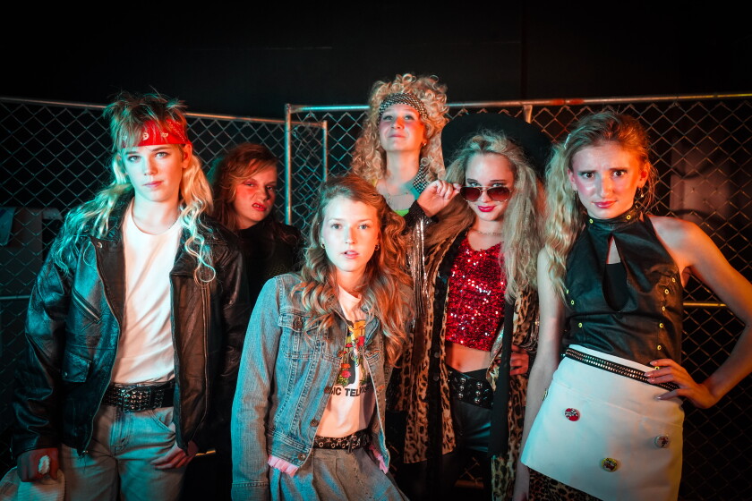 Seis adolescentes de pele clara, vestidos com roupas de estrela do rock dos anos 80, posam para uma foto em grupo no pano de fundo de uma cerca de arame.