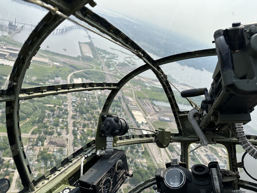 View from the nose of a B-25 World War II bomber. In the foreground, a window bubble and machine gun controls are visible. In the background, a river and cityscape.