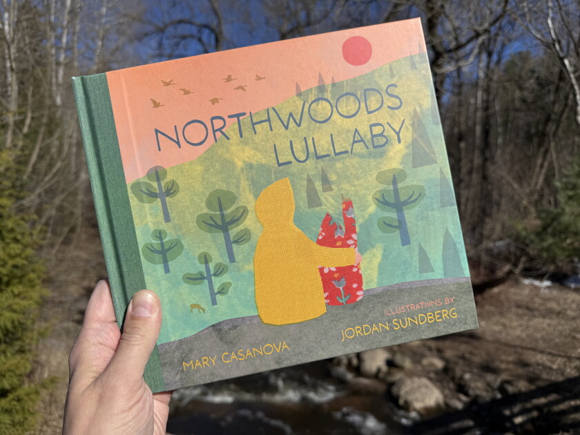 White man's hand holds picture book: "Northwoods Lullaby" by Mary Casanova, with illustrations by Jordan Sundberg. Cover illustration depicts adult and child sitting together, wearing hooded jackets.