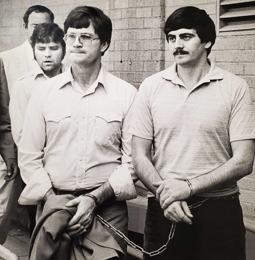 Two men in the foreground with hands cuffed in front of them and a chain connecting them. Another is in the background.