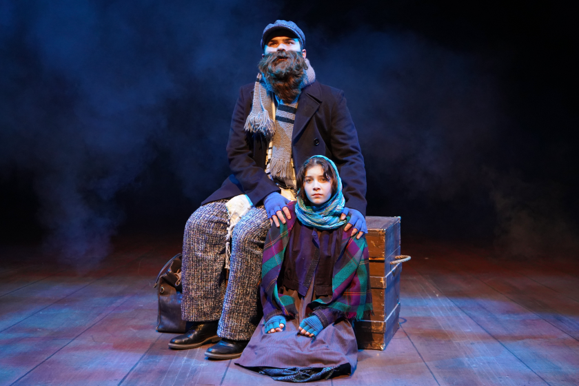 A man and young girl, dressed in humble but warm clothing, huddle together with a steamer trunk.