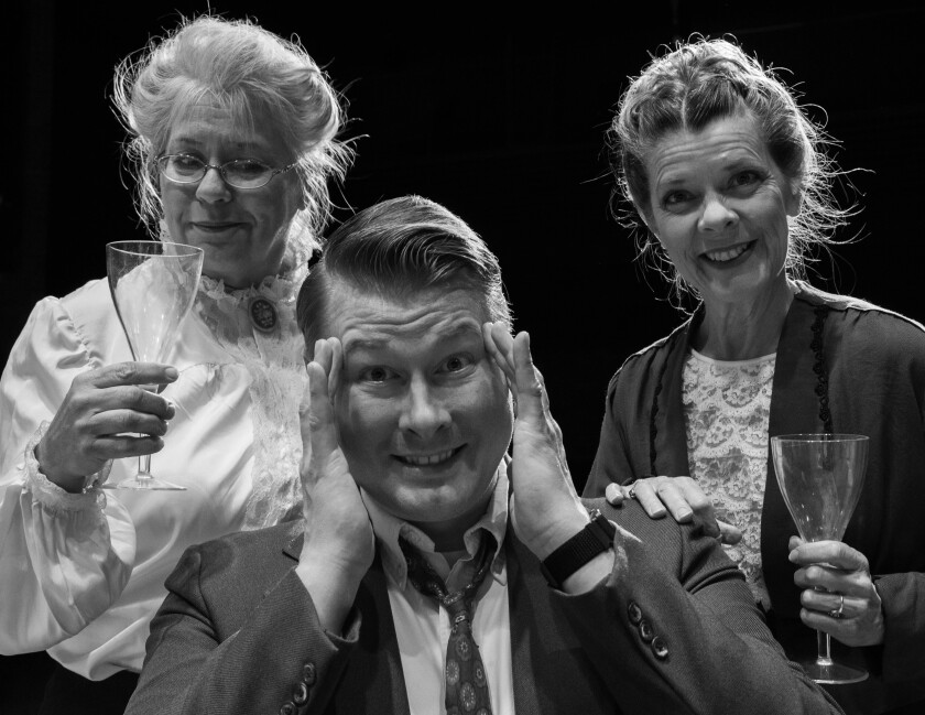Black and white photo of three people posing in vintage garb indicating an 1940s setting. Two women smile and hold empty glasses on either side, while a man in the center puts his hands to his temples and half-smiles, half-grimaces.