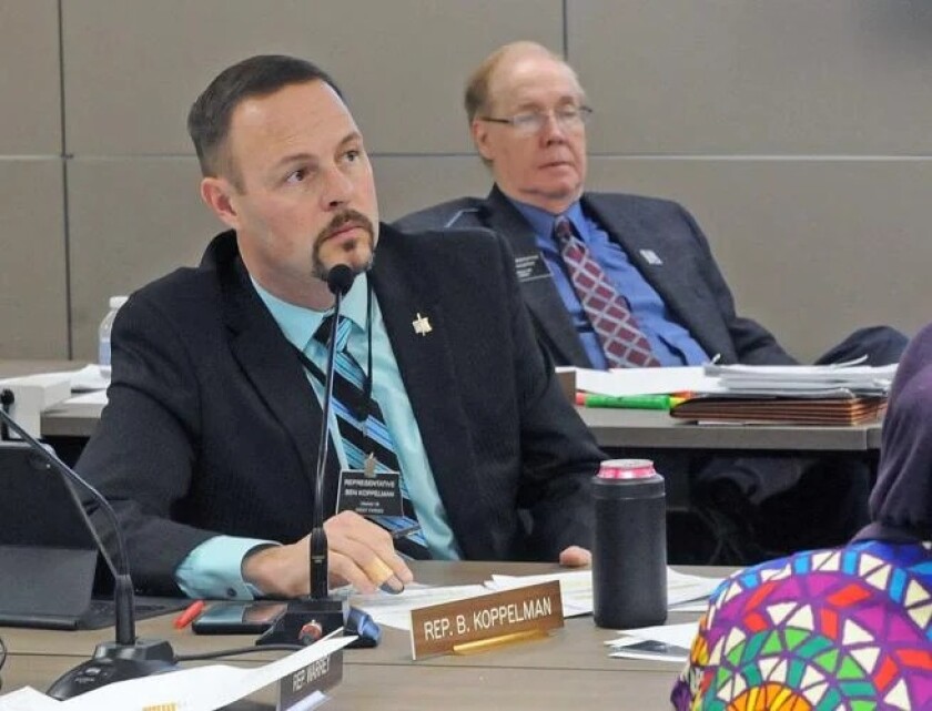 A goateed man in a black suit coat, light blue shirt, and blue and black striped tie looks to the side while seated in front of a name plate and microphone. The name plate reads "Rep. B. Koppelman."