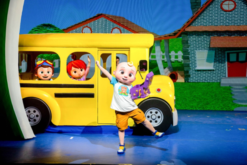 A performer in a giant baby head dances in front of a yellow school bus in a staged show.