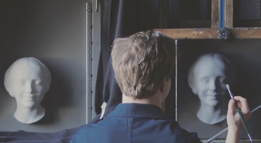 A painter, seen from behind, works to capture the nuances of light and shadow on a bust he's rendering on canvas.