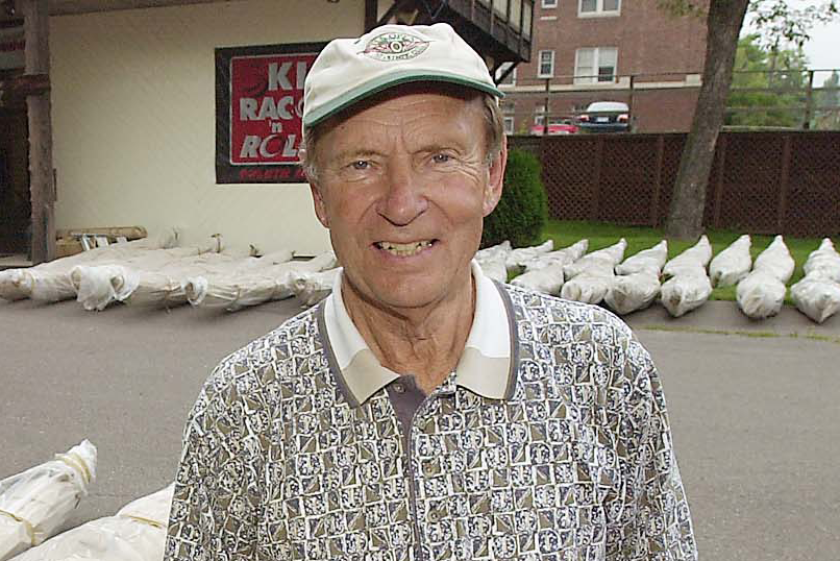 Elderly man in a white cap stands in front of kayaks wrapped in plastic.