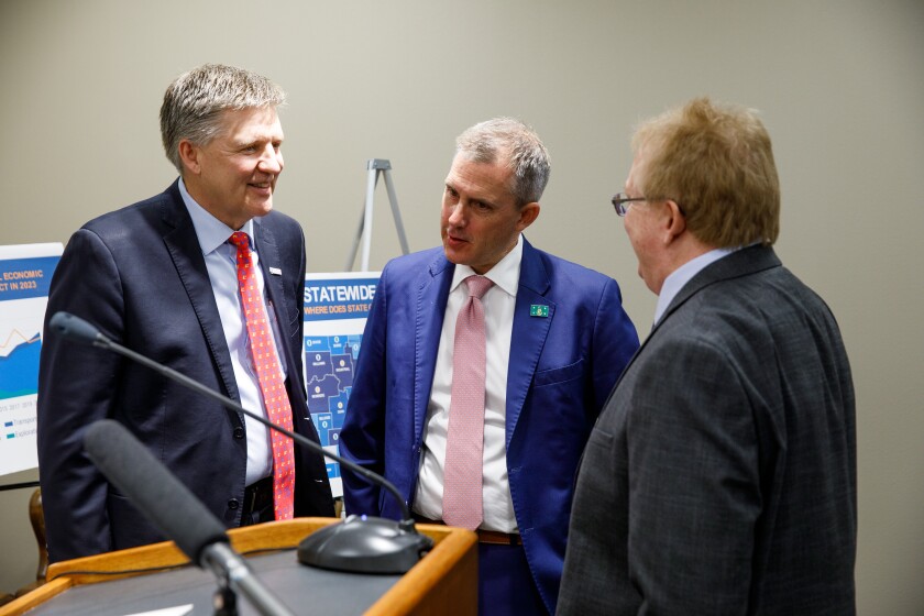 Three mens in suits and ties talk together next to a podium with several microphones