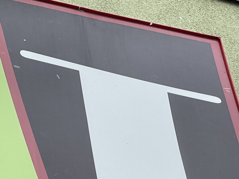 Part of a stylized letter - possibly an I, J or T - on a panel, printed in white on black with a visible red and green border around the edges.