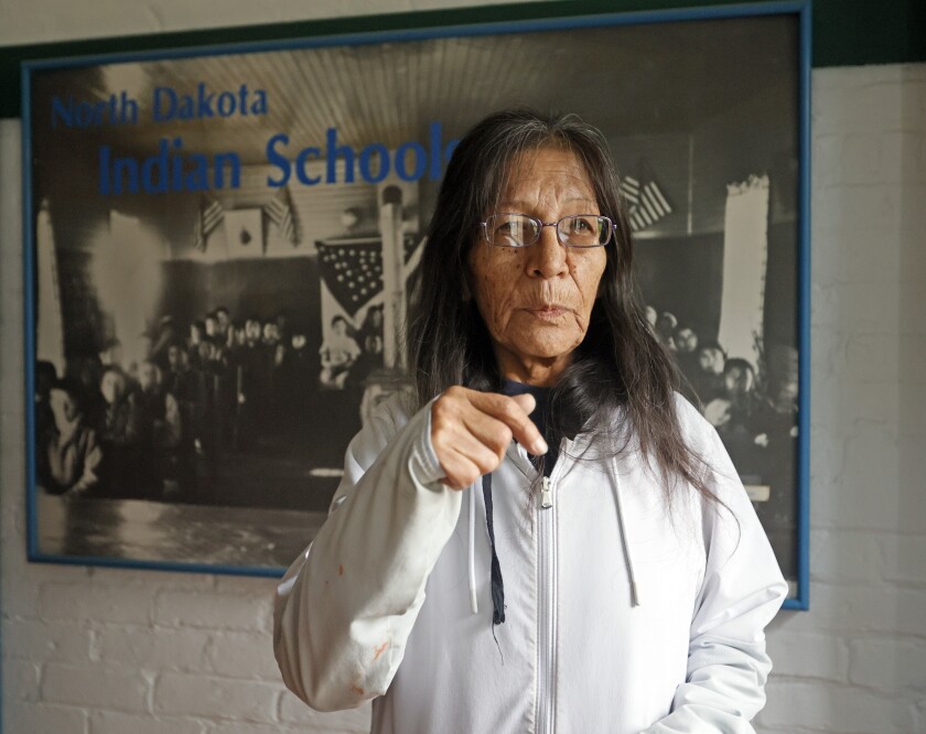 A woman in a white jacket stands in front of a black and white poster with the blue words "North Dakota Indian Schools" on it.