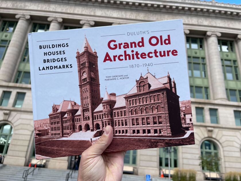 Hand holding book, "Duluth's Grand Old Architecture 1870-1940: Buildings, Houses, Bridges, Landmarks" in front of a tall neoclassical stone building.