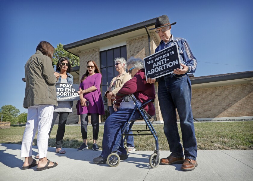 Several women, one sitting on a walker and a man stand outside an office building with anti-abortion signs.