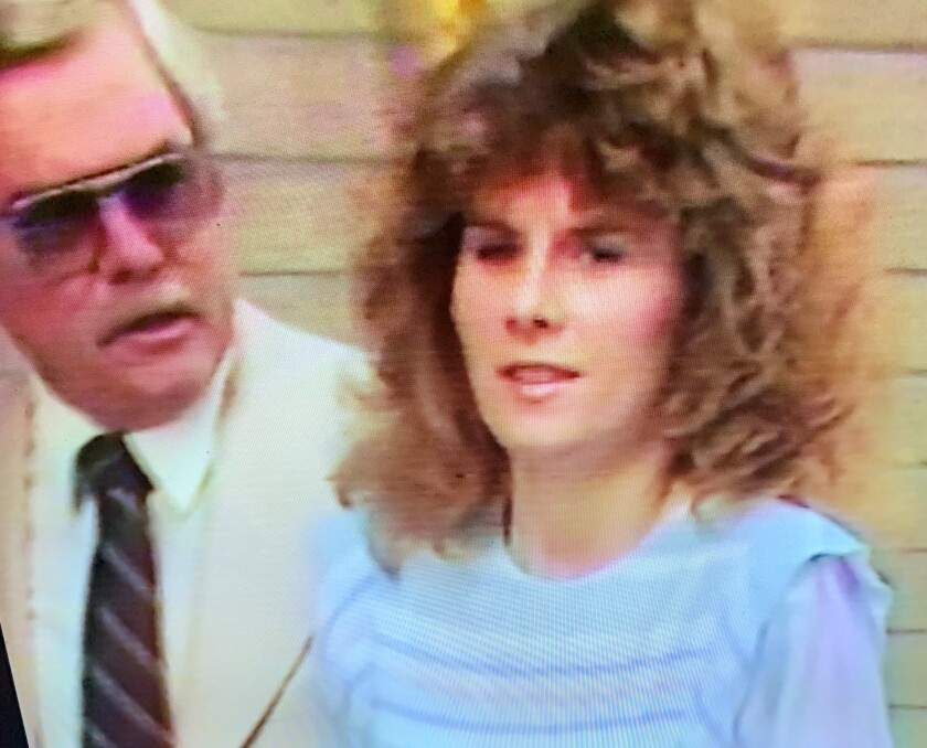 A woman with curly brown hair stands in the foreground. A mustached man with sunglasses stands in the background.