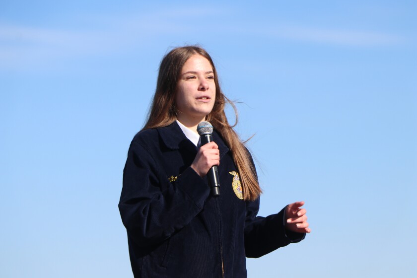 A brown-haired high school FFA member addresses a crowd of 300, flanked by the blue sky.