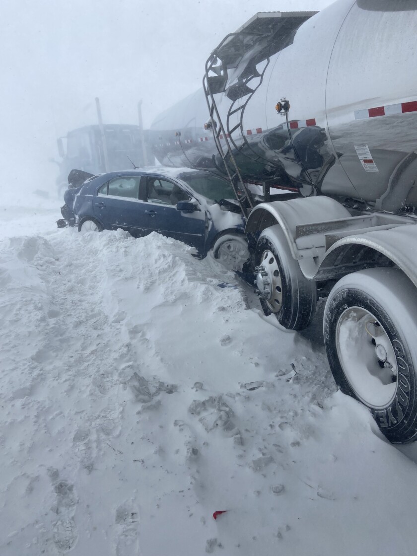 The hood of a small blue sedan is partially crushed under a tank being pulled by a semi. The back of the sedan is crushed inward.