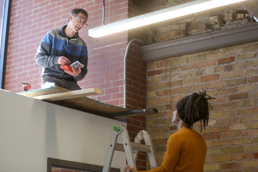 A man sitting above an office while another man stands on a ladder.