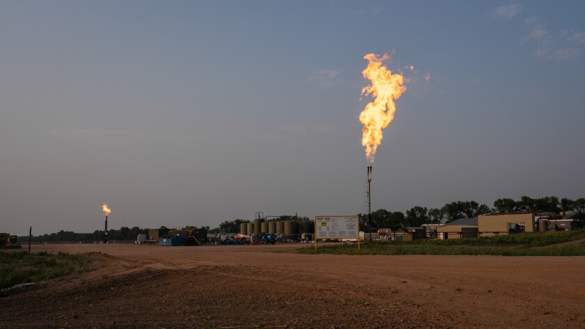 flames erupt out of the top of a large metal pipe at an industrial site with low buildings and tanks