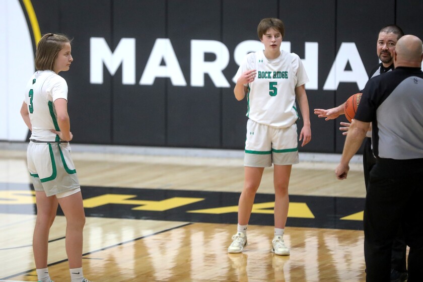Two high school girls basketball players looking on while officials chat during a break in the action of a game.