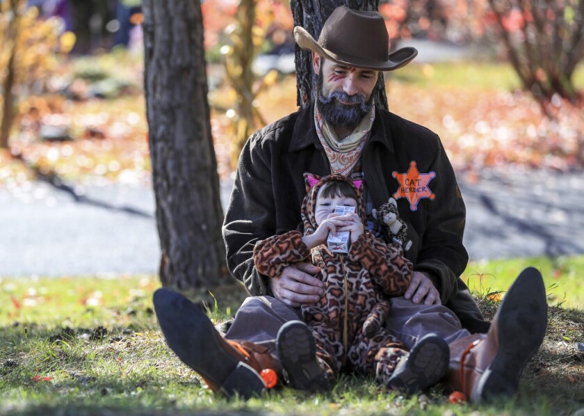 A man dressed as a sheriff and a young girl dressed as a cat sit on a grassy slope while the child drinks juice. Fall leaves are visible out of focus in the background.