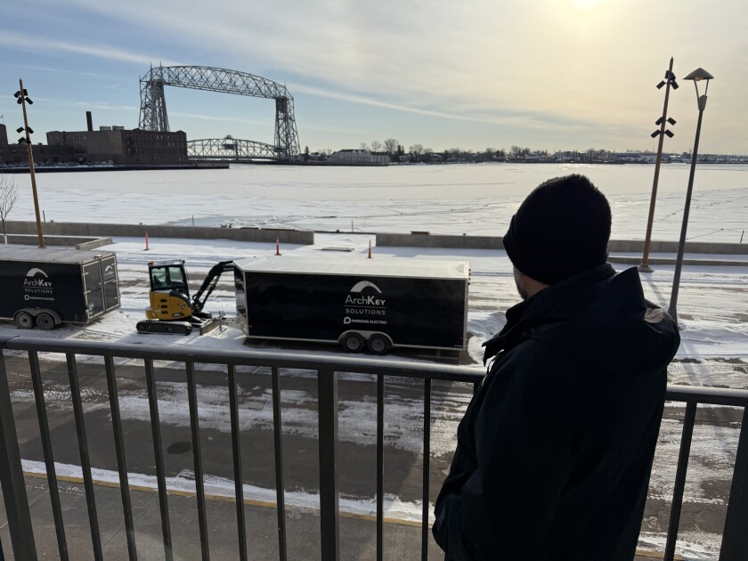 Man in black jacket and winter hat is seen from behind, looking over lake harbor with lift bridge in background. Railing stands behind him.