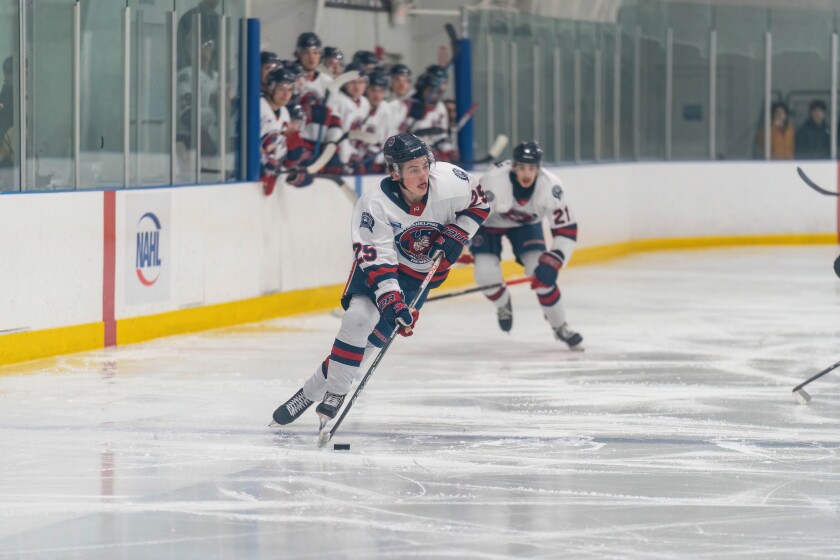 A hockey player wearing a dark blue, red, and white jersey skates up the ice with the puck.