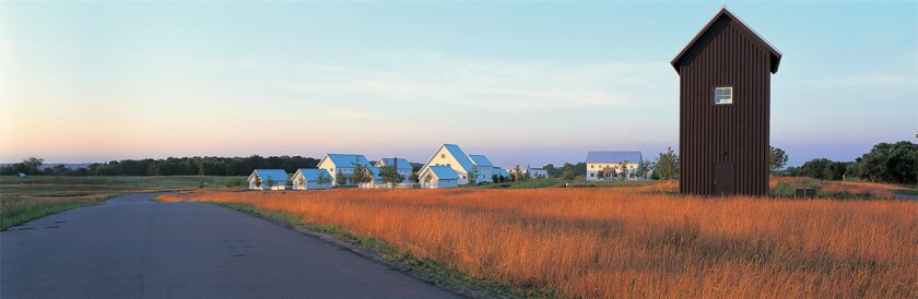 Several white houses, in background, and a tall but narrow brown building, at right foreground, sit in a meadow as seen from a smooth roadway at dusk.